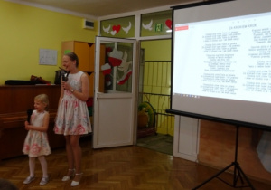 Dziewczynka z mamą śpiewa piosenkę, na ekranie wyświetlane są słowa piosenki.